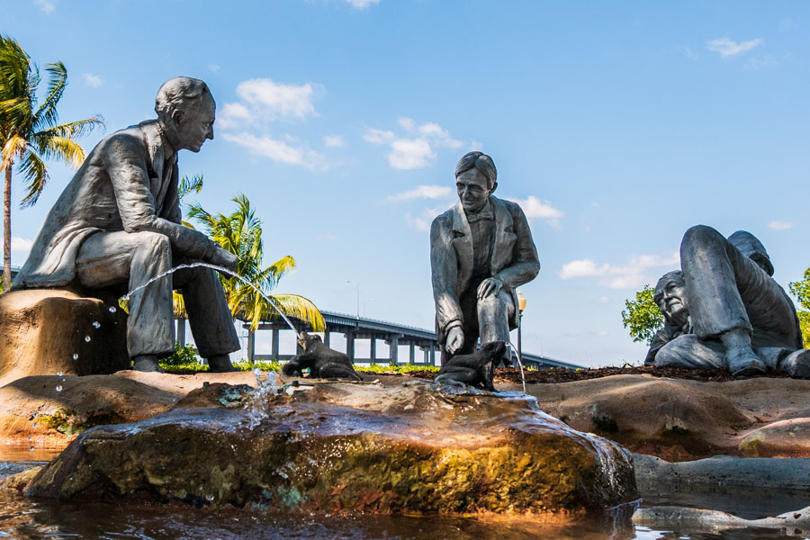 Henry Ford sitting, Harvey Firestone kneeling and Thomas Edison reclining around a campfire on an island within a fountain. 