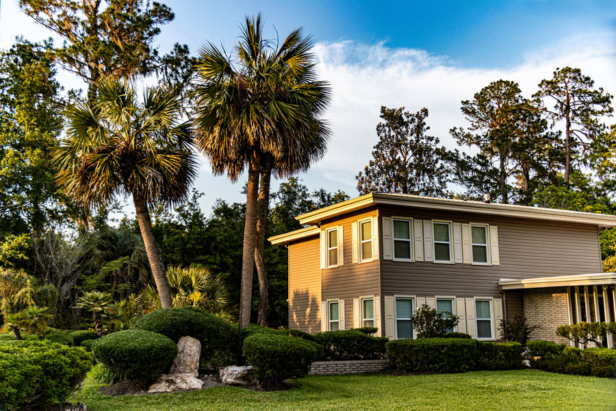 A two-story mid-century modern home sits in a landscaped setting of sculptured shrubs, palm trees, picturesque rocks and a manicured green lawn. 